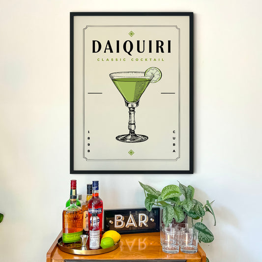 a framed poster of a Daiquiri cocktail hanging on a wall above a bar counter with various liquor bottles and glasses.