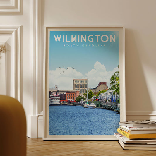 A framed poster of a cityscape with a river in the foreground, displaying the text "WILMINGTON NORTH CAROLINA" and a few boats on the water.