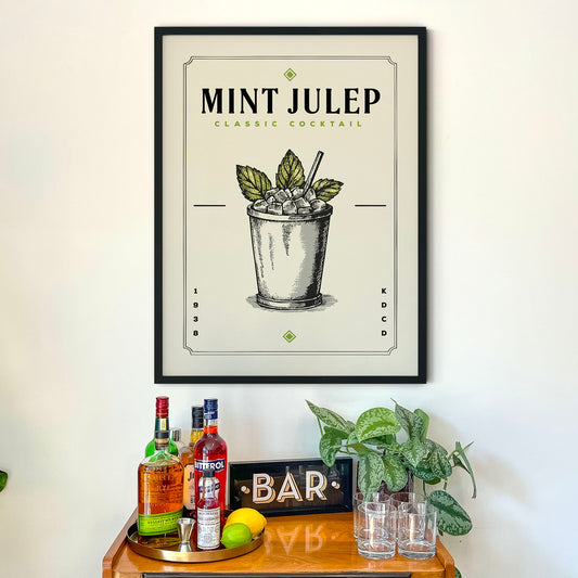 a framed poster of a mint julep cocktail hanging on a wall above a bar cart. The poster includes illustrations of the cocktail and the word "MINT JULEP" prominently displayed.