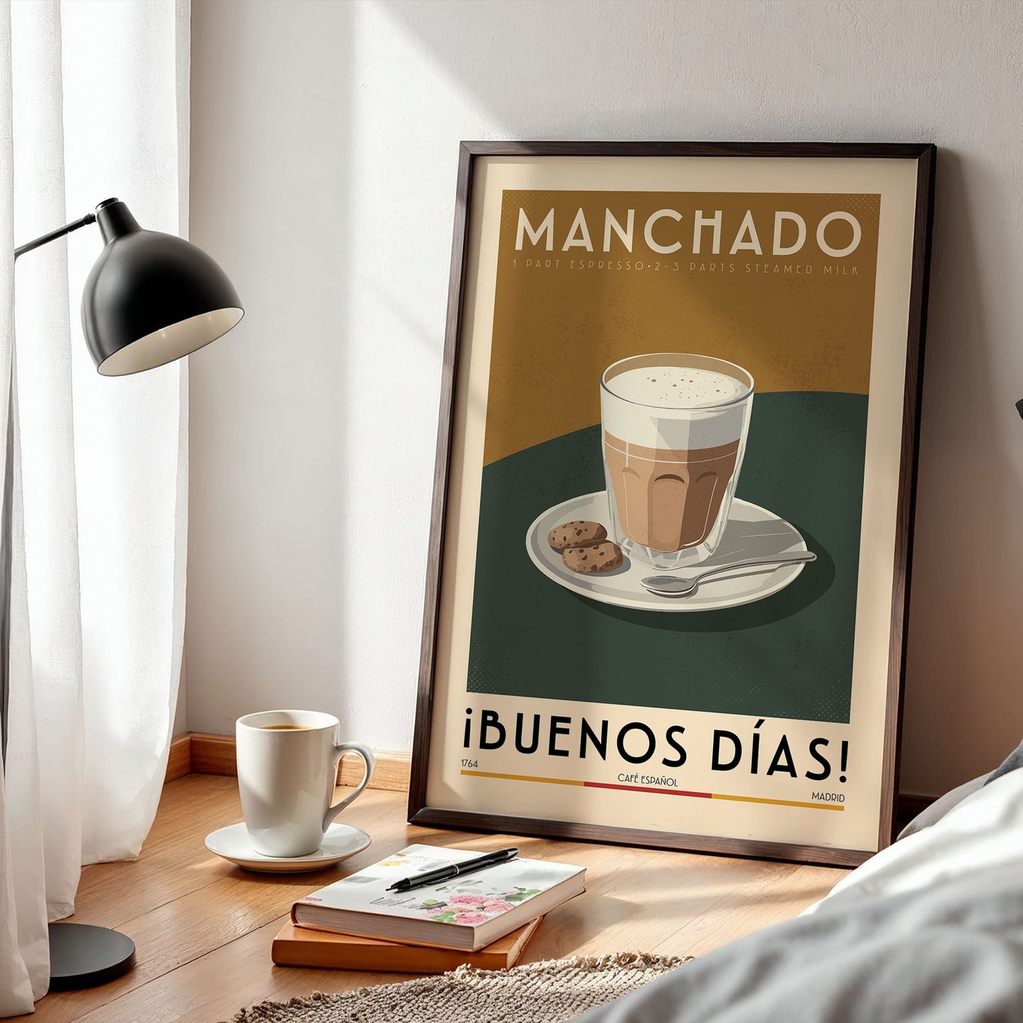 a framed poster of a cup of coffee with the text "Manchado" and "¡Buenos dias!" (Good Morning!) written on it, along with an illustration of a cup of coffee and a plate of cookies. The poster is displayed on a wooden table, and there is a lamp, a book, and a cup of coffee nearby.