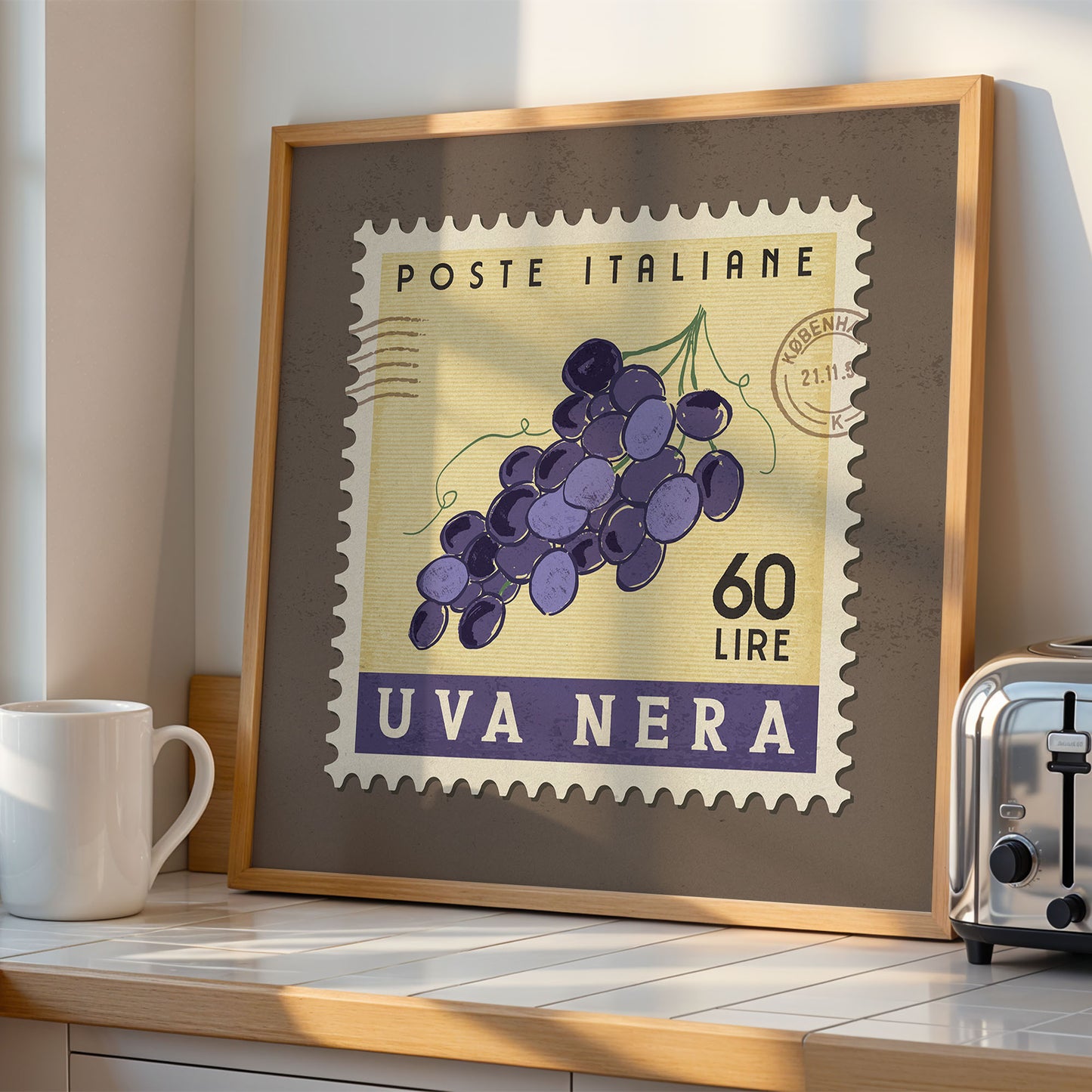 a framed stamp-like artwork depicting a bunch of purple grapes, with the text "POSTE ITALIANE 60 LIRE UVA NERA" printed on it. The artwork is displayed on a shelf next to a toaster, and there is a white mug visible in the background.