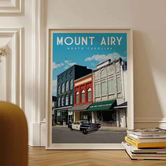 A framed poster of a street scene in Mount Airy, North Carolina, with a vintage car parked on the street and colorful buildings in the background.