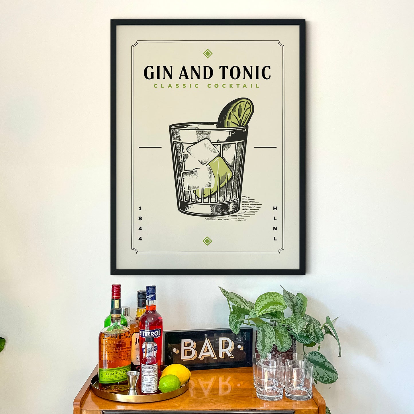 a framed poster of a gin and tonic cocktail hanging on a wall above a wooden bar cart. The poster displays an illustration of a gin and tonic glass with ice cubes and a lime wedge. The bar cart has various bottles of alcohol and glasses on it, suggesting a bar setting.
