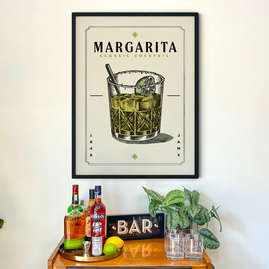 a framed poster of a Margarita cocktail hanging on a wall above a wooden bar cart. The poster displays the Margarita cocktail illustration and includes the text "MARGARITA CLASSIC COCKTAIL" and "SAR" below the illustration. The bar cart is filled with various liquor bottles and has a sign that reads "BAR".