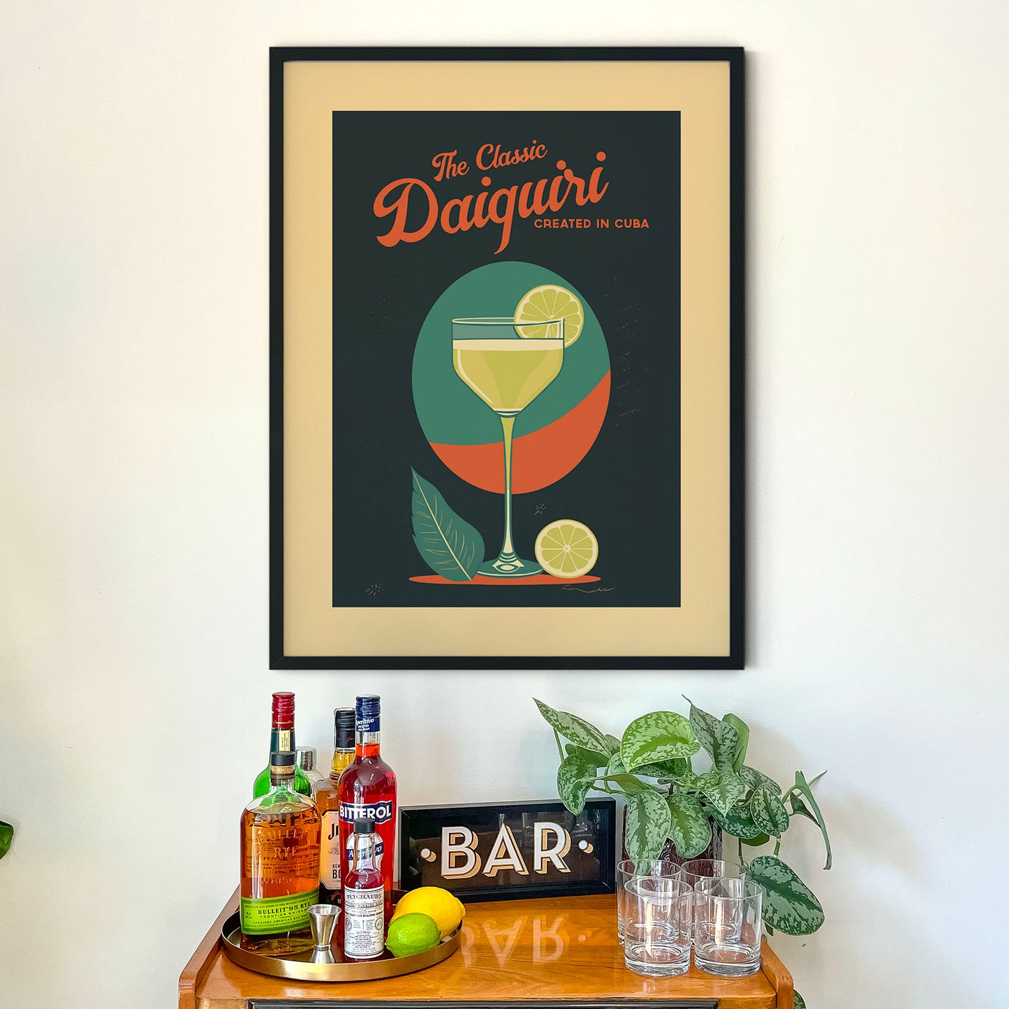 a framed poster of a classic daiquiri cocktail, with a glass of daiquiri and various ingredients such as a lemon and a lime. Below the poster, there is a wooden bar cart with several bottles of alcohol and a potted plant.