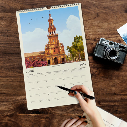 A person's hand is holding a pen and writing on a calendar, with a camera and other items on a wooden surface.