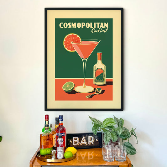 a framed poster of a Cosmopolitan cocktail on a wall above a bar cart with various liquor bottles and a potted plant.