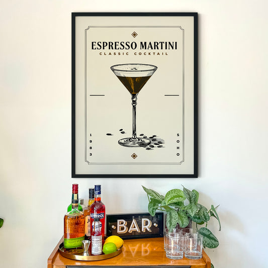 a framed poster of an espresso martini cocktail hanging on a wall above a wooden bar cart. The poster displays an illustration of the cocktail glass and the word "ESPRESSO MARTINI" along with the text "CLASSIC COCKTAIL". The bar cart is filled with various liquor bottles and has a sign that reads "BAR".