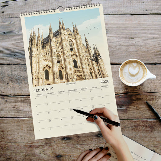 A person's hand is holding a pen and writing on a calendar page featuring a photograph of a cathedral, with a cup of coffee and a notebook on a wooden surface in the background.