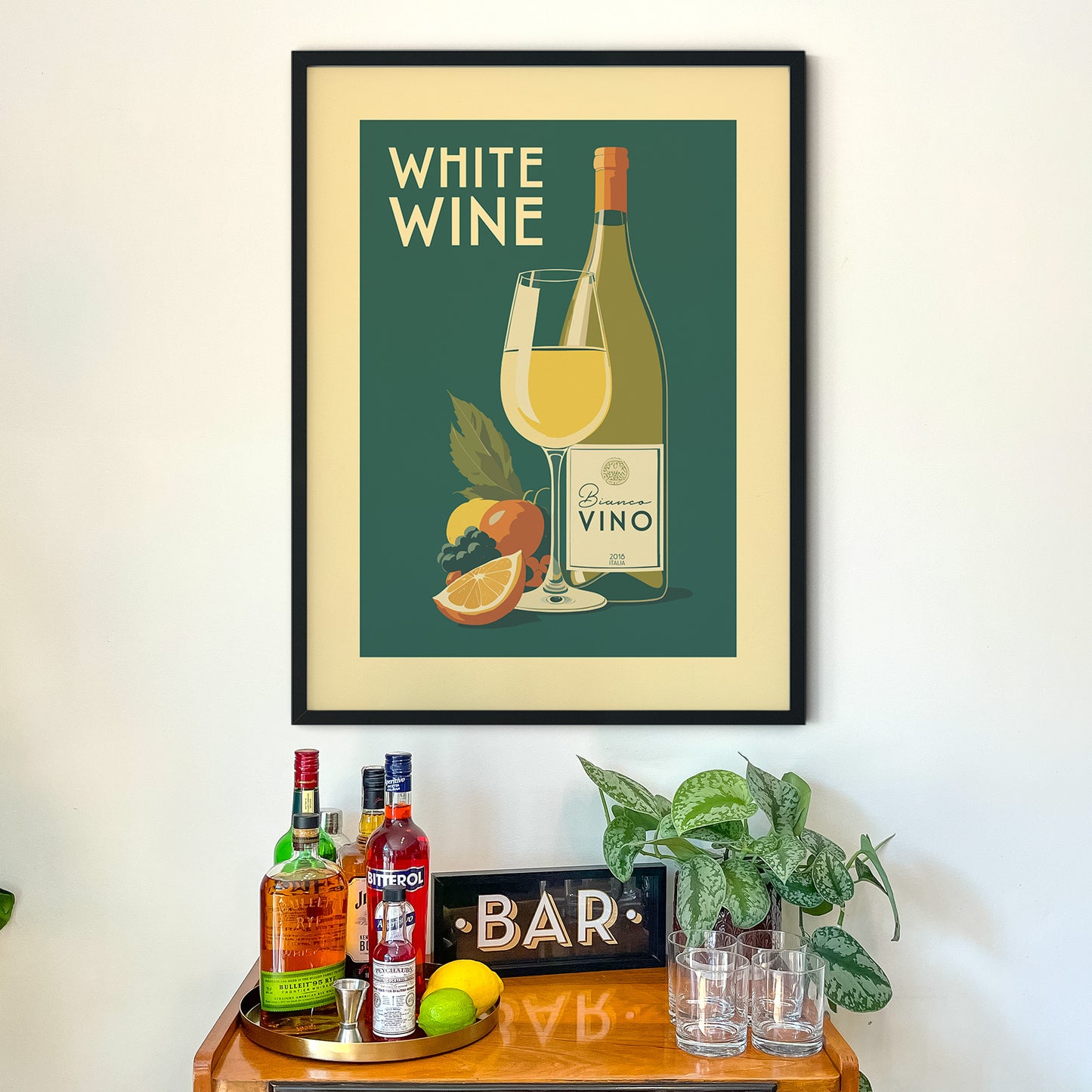 a framed poster of a wine bottle and glass, along with various bottles of alcohol and a potted plant, all displayed on a wooden table.