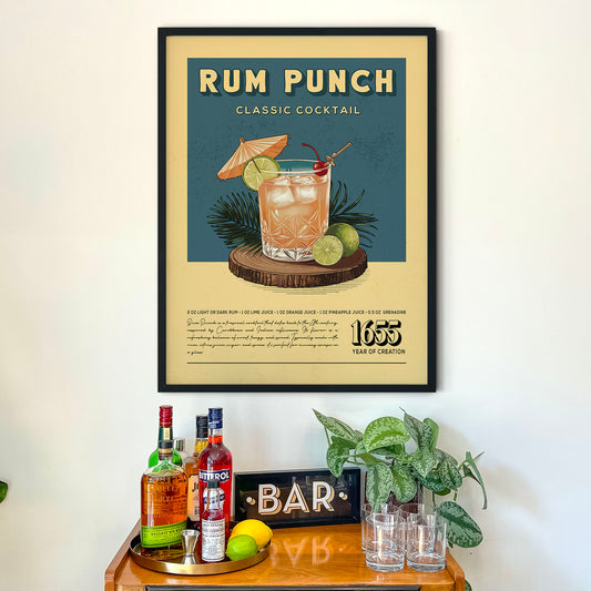 a framed poster of a classic cocktail called "RUM PUNCH" with illustrations of the drink and garnishes, such as a lime and an umbrella, displayed on a wall above a wooden bar counter with various liquor bottles and glasses.