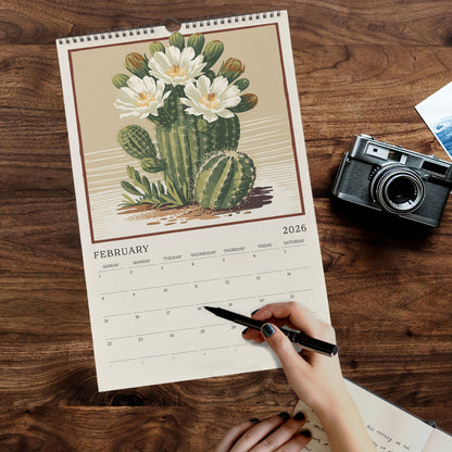 A person's hand is writing in a notebook while holding a pen, with a calendar featuring a cactus illustration on a wooden surface nearby.