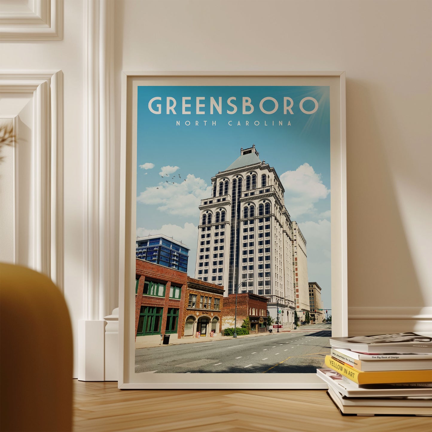 A framed poster of Greensboro, North Carolina, featuring a tall white building with a blue roof against a blue sky with clouds.