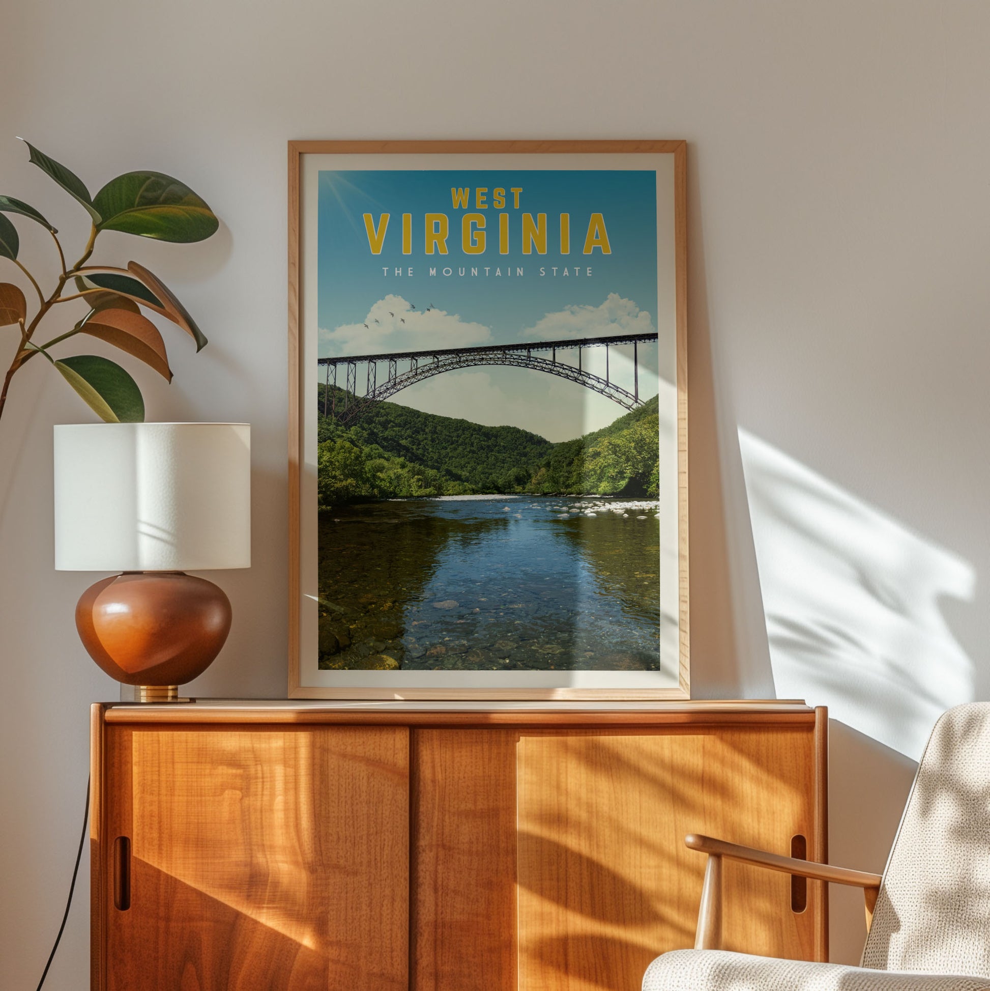 a framed poster of a bridge over a river, with the text "West Virginia" and "The Mountain State" written on it. The poster is displayed on a wooden cabinet, and there is a lamp and a plant in the foreground.