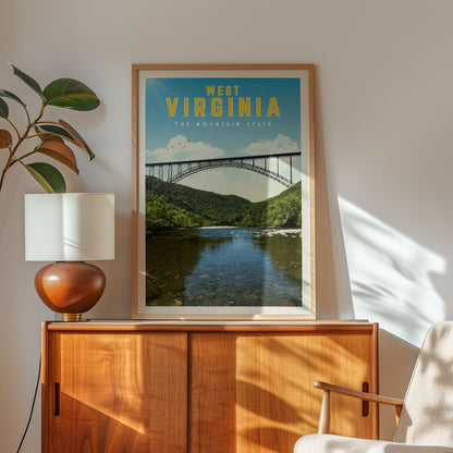 a framed poster of a bridge over a river, with the text "West Virginia" and "The Mountain State" written on it. The poster is displayed on a wooden cabinet, and there is a lamp and a plant in the foreground.