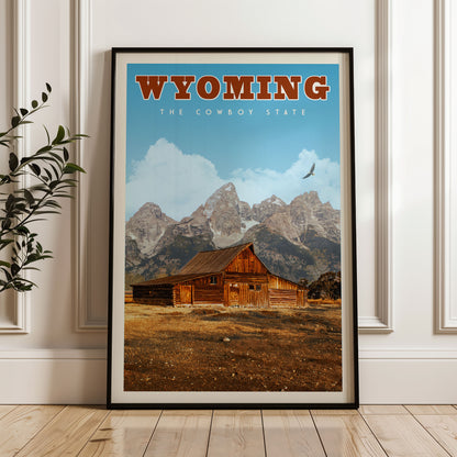 A framed poster of a rustic wooden cabin set against a backdrop of majestic mountains, with the text "Wyoming The Cowboy State" prominently displayed.
