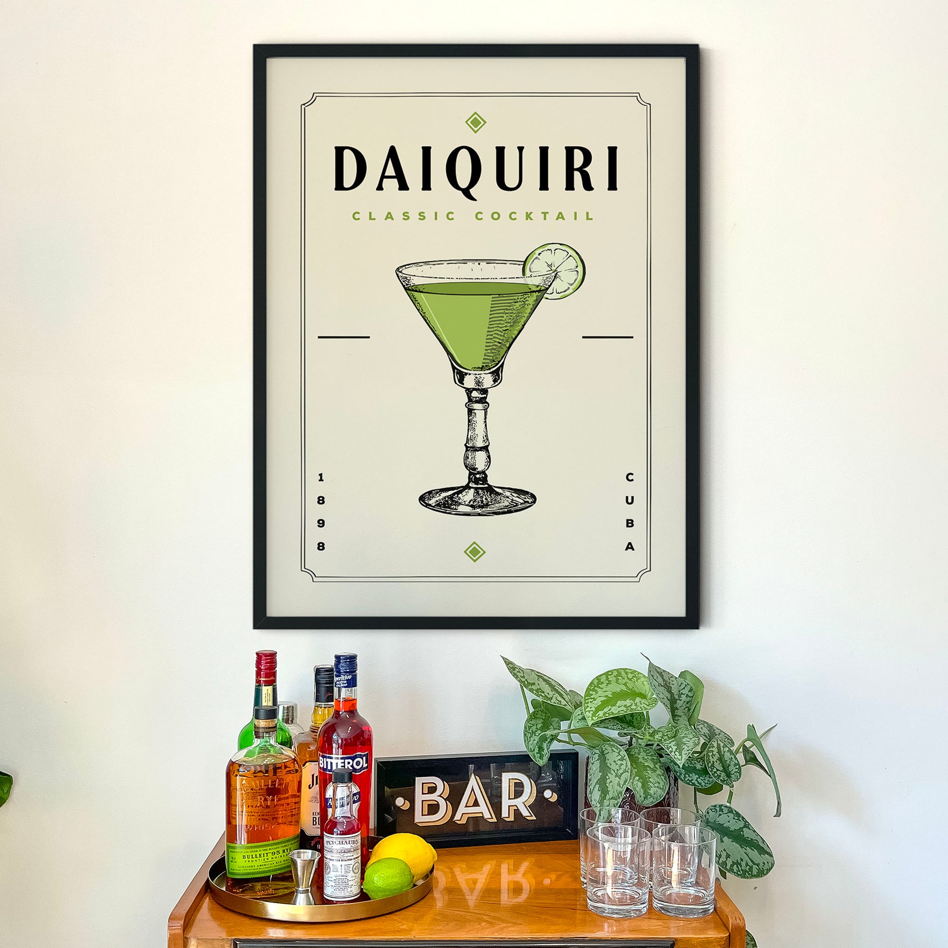 a framed poster of a Daiquiri cocktail hanging on a wall above a bar counter with various liquor bottles and glasses.