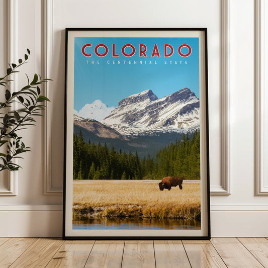 A framed poster of a scenic landscape in Colorado, featuring a bison grazing in a field with snow-capped mountains in the background.