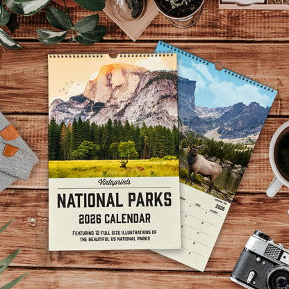 a calendar with a scenic mountain landscape on the cover, and a smaller calendar with a photo of a deer on the cover. The calendar is titled "Vintoprints NATIONAL PARKS 2026 CALENDAR" and includes a feature of 12 full-size illustrations of the beautiful US national parks. The smaller calendar also has a photo of a deer on the cover. The background includes a wooden table with various items such as a cup, a plant, and a camera.