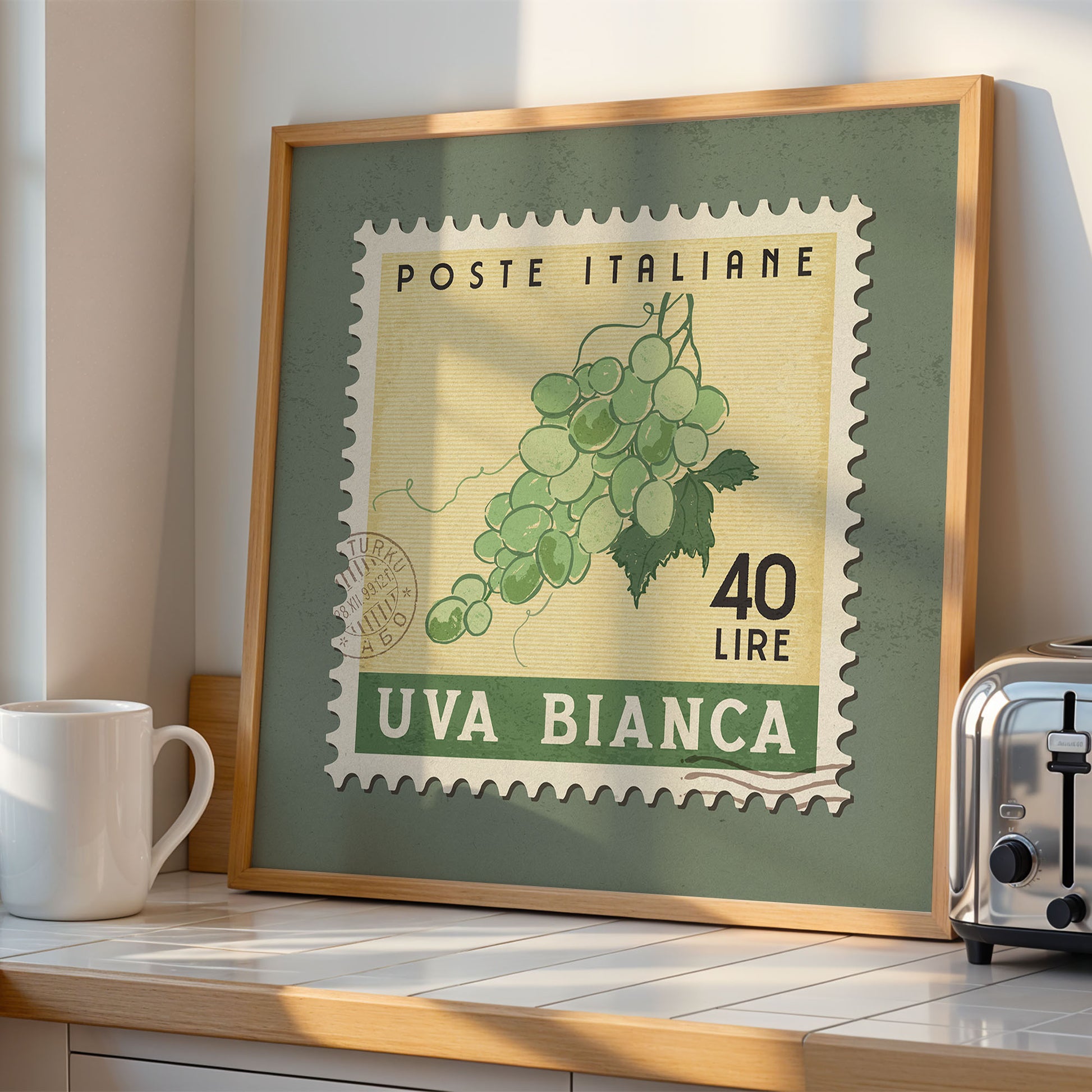 a framed Italian postage stamp with a green background and a depiction of grapes. The stamp is labeled "POSTE ITALIANE" and "40 LIRE", and it is displayed on a kitchen counter. There is also a white coffee mug and a toaster visible in the scene.