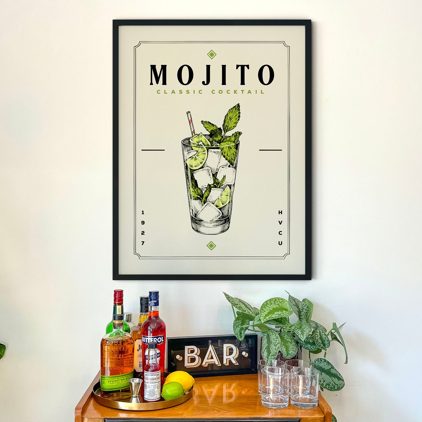 a framed poster of a mojito cocktail hanging on a wall above a wooden bar cart. The poster displays the mojito cocktail illustration and includes text that reads "MOJITO CLASSIC COCKTAIL". Below the poster, there are various bottles of alcohol and glasses on the bar cart, suggesting a bar setting.
