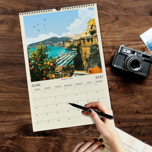 A person's hand is holding a pen and writing on a calendar page that features a scenic view of a coastal town with a harbor and mountains in the background.