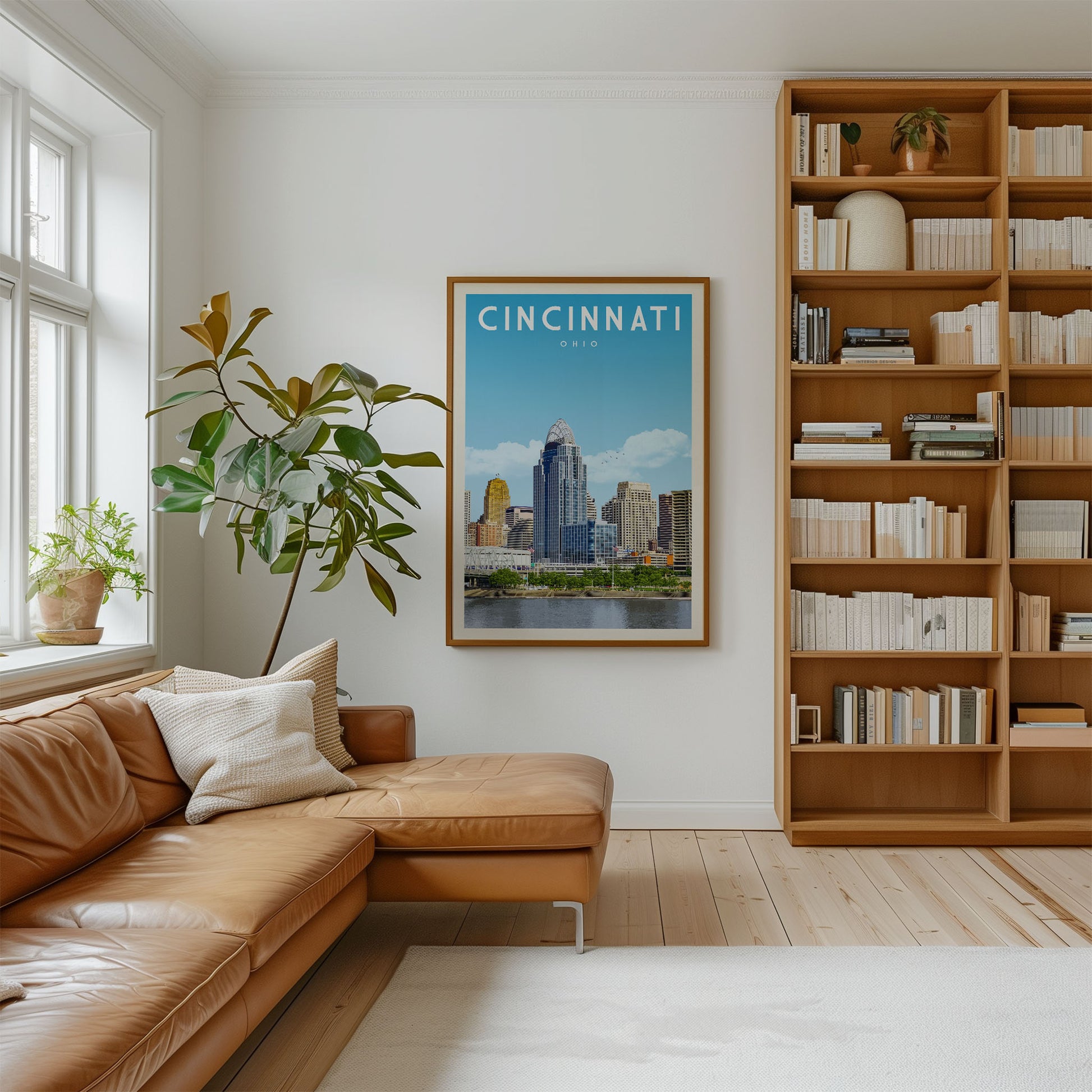 a cozy living room with a large window, a comfortable leather couch, a bookshelf filled with books, and a framed poster of the Cincinnati skyline hanging on the wall.
