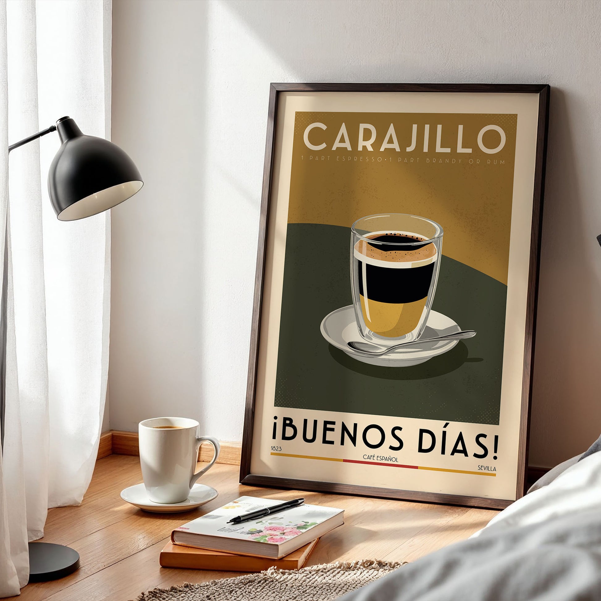 a framed poster of a cup of coffee with the text "CARAJILLO" and "¡Buenos dias!" (Good Morning!) on it, placed on a wooden floor next to a lamp and a book.