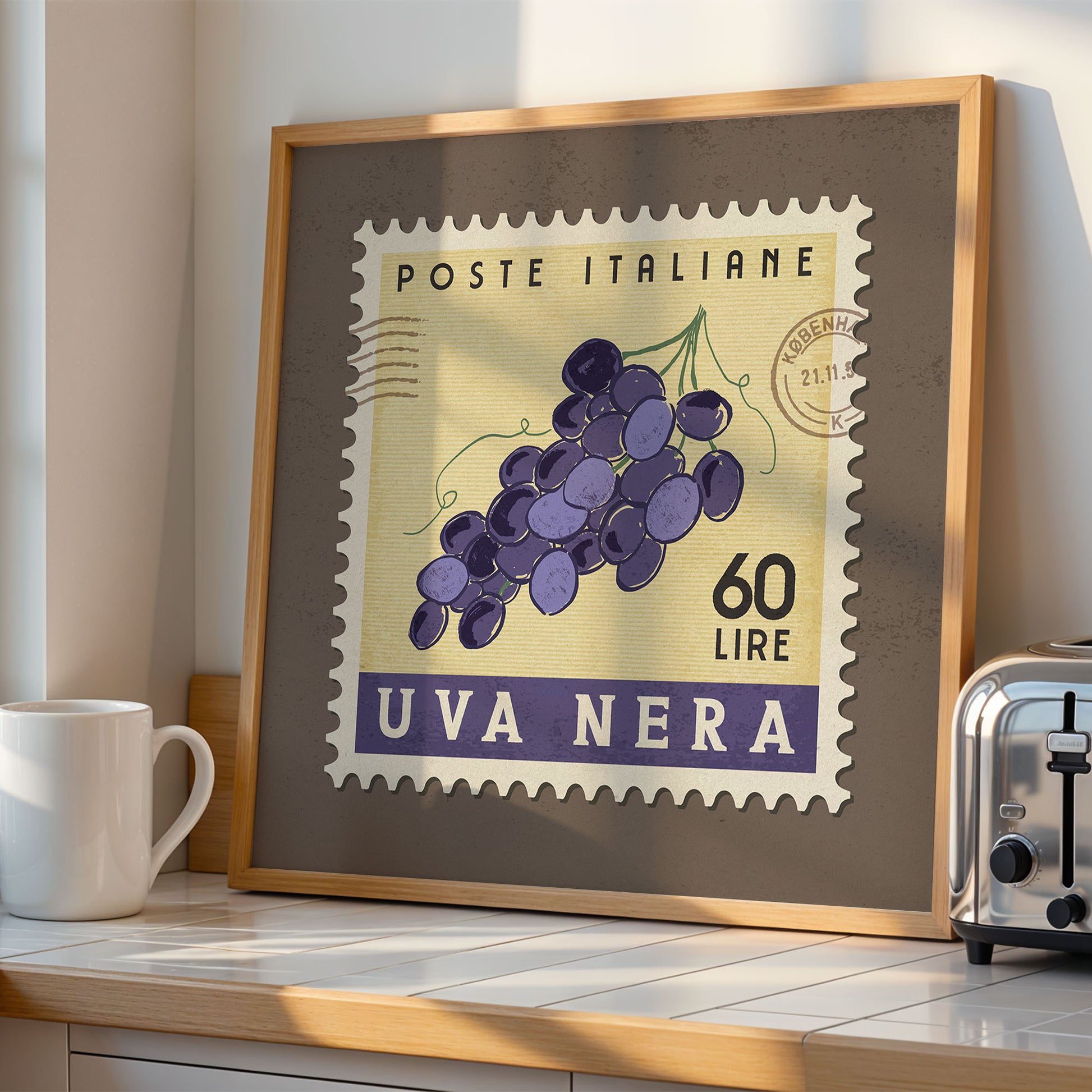 a framed stamp-like artwork depicting a bunch of purple grapes, with the text "POSTE ITALIANE 60 LIRE UVA NERA" printed on it. The artwork is displayed on a shelf next to a toaster, and there is a white mug visible in the background.