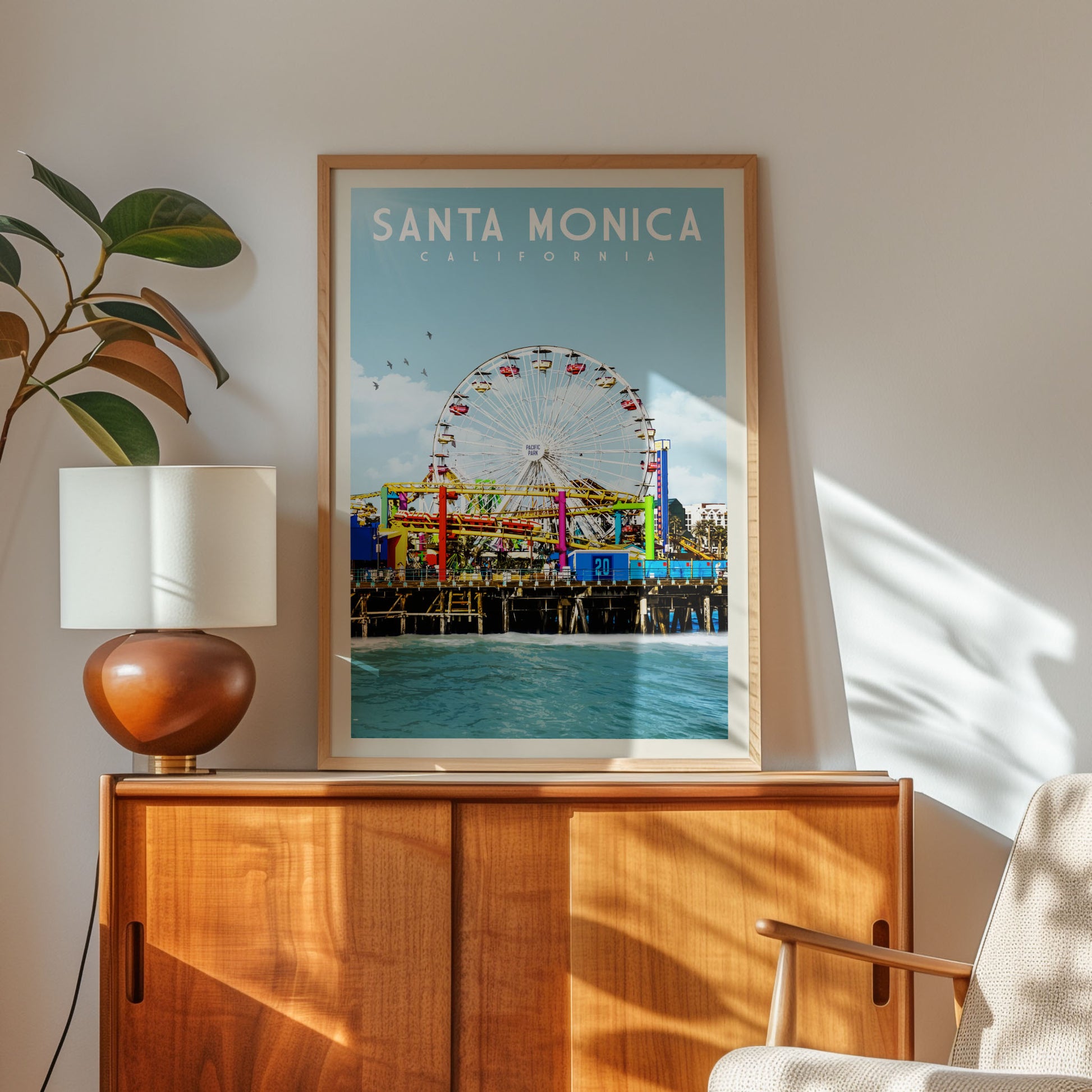 a framed poster of the iconic Santa Monica Ferris wheel in California, with a wooden cabinet and a plant in the foreground.