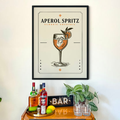 a framed poster of an Aperol Spritz cocktail hanging on a wall above a wooden bar cart. The poster displays the cocktail illustration and the text "Aperol Spritz Classic Cocktail".