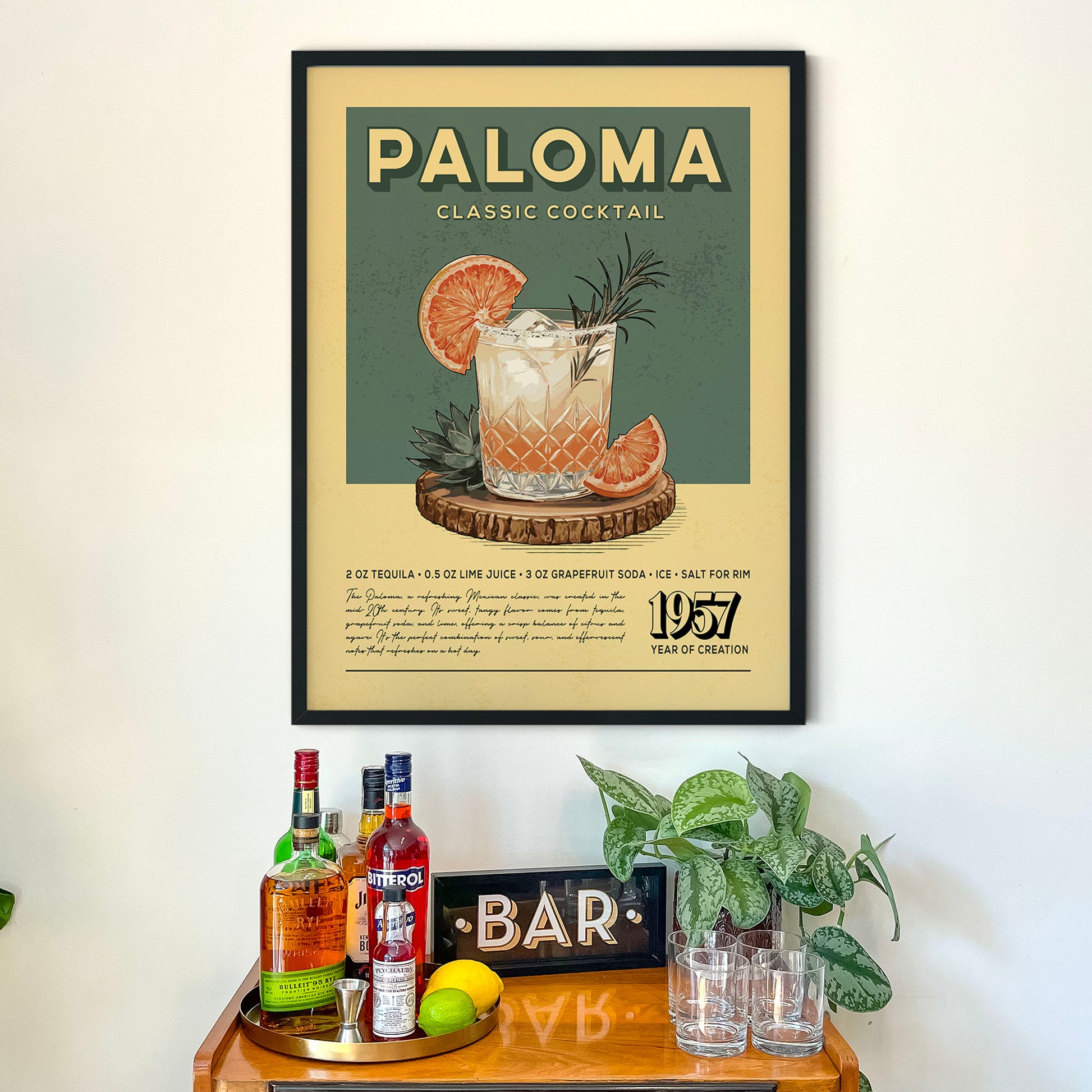 a framed poster of a cocktail called "Paloma Classic Cocktail" hanging on a wall above a wooden bar display with various liquor bottles and glasses.