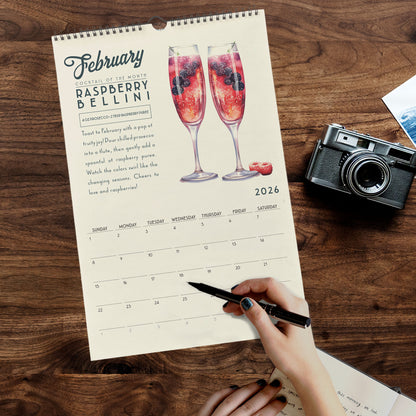 A person's hand is writing in a notebook while holding a pen, with a calendar for February on a wooden surface in front of them.