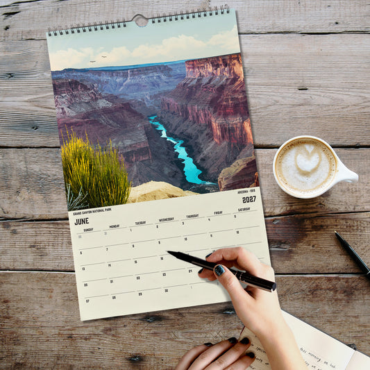A person's hand is writing in a notebook while holding a pen, with a coffee cup and a scenic view of the Grand Canyon in the background.