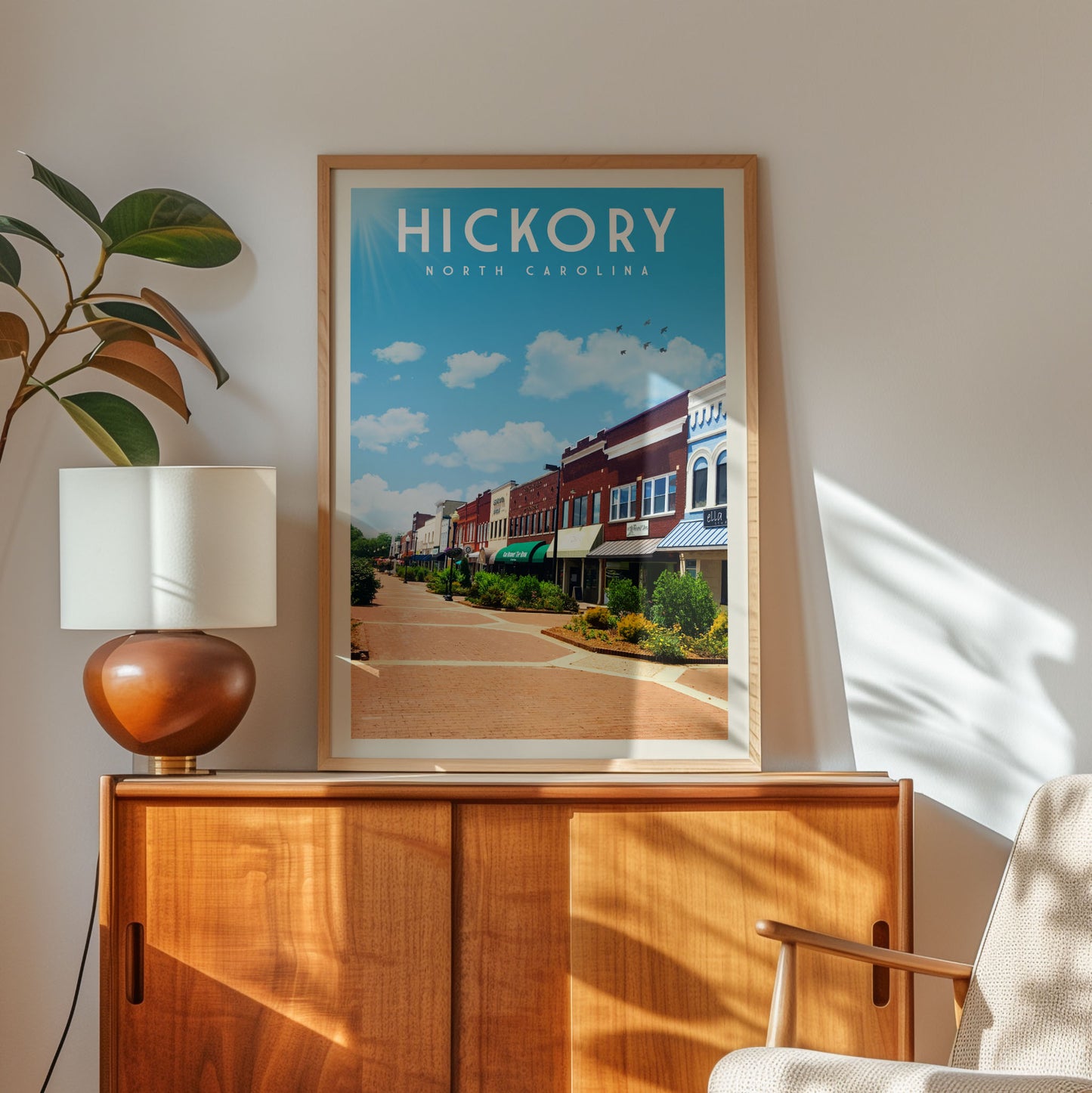 a framed poster of a street scene in Hickory, North Carolina, hanging on a wall next to a wooden cabinet and a lamp.
