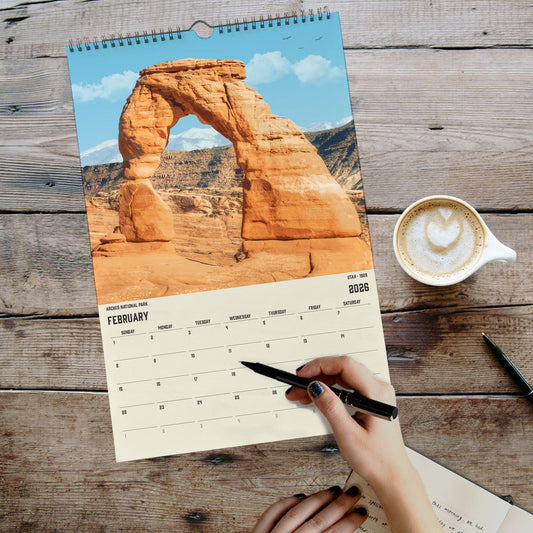 A person's hand is writing in a notebook while holding a pen, with a calendar featuring a desert landscape and a cup of coffee on a wooden surface.