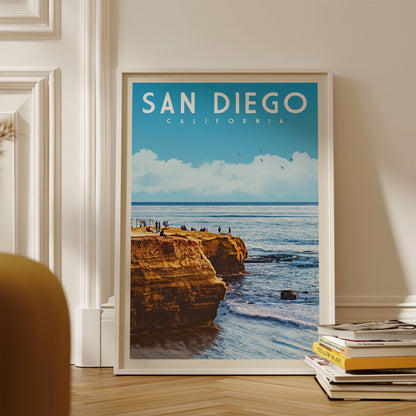 A framed poster of a scenic coastal landscape with cliffs and people enjoying the view, displayed on a wall next to a stack of books.