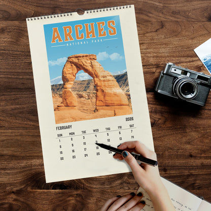 A person is writing in a notebook while holding a pen, with a calendar featuring a scenic image of a natural arch in the background.
