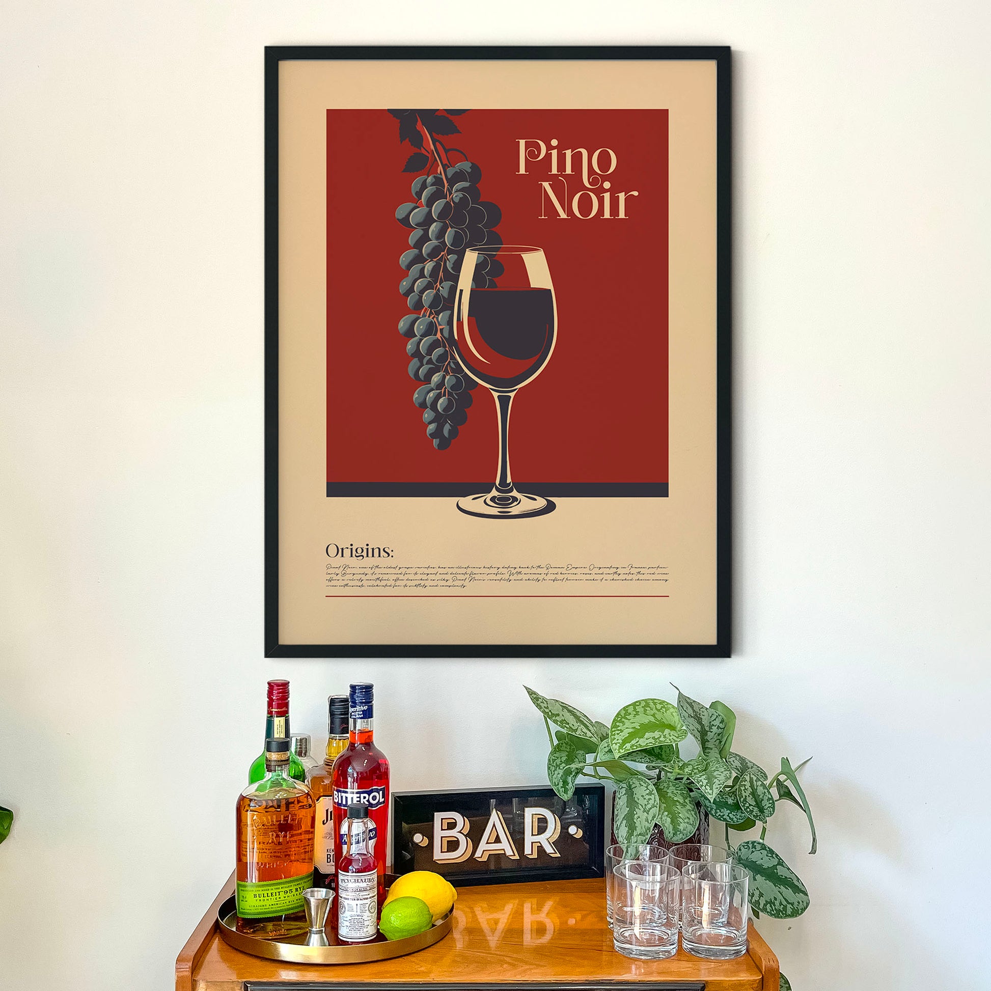 a framed poster of a wine glass with grapes hanging from it, placed above a wooden bar counter with various bottles of alcohol and a potted plant.
