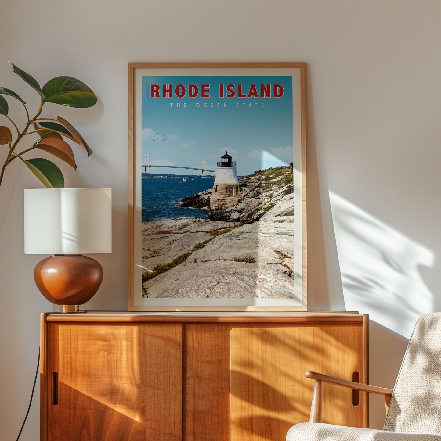 a framed poster of a lighthouse on a rocky coastline, with a bridge in the background, hanging on a wall next to a wooden cabinet and a lamp.