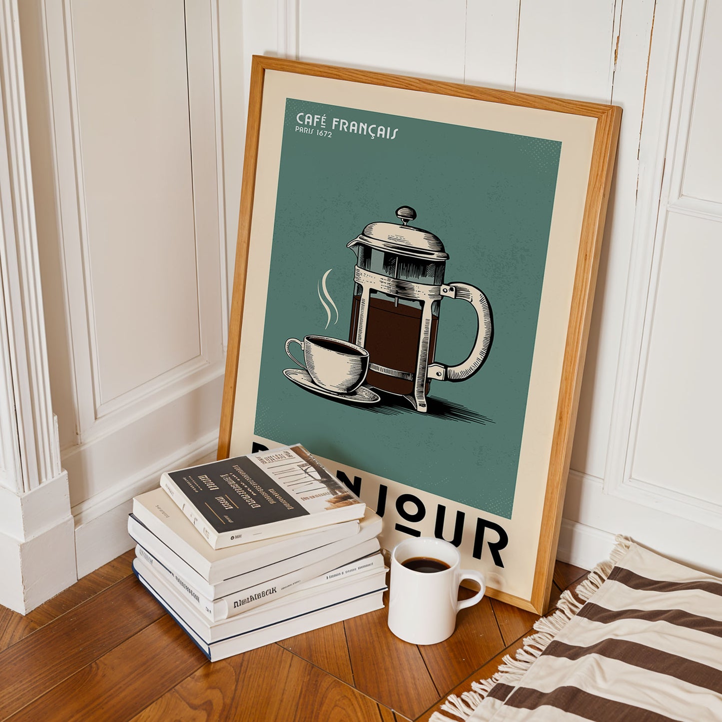 a framed poster of a French press coffee maker, a stack of books, and a white mug on a wooden floor.