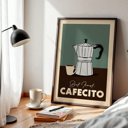A framed poster of a coffee maker is displayed on a wooden floor next to a lamp, a cup of coffee, and some books.