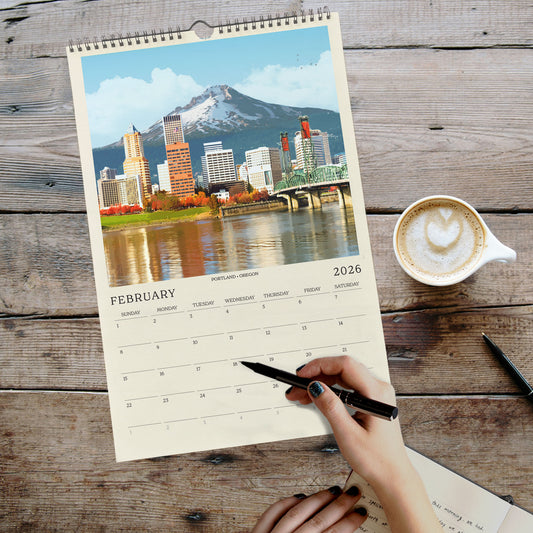 A person's hand is writing in a notebook while holding a pen, with a coffee cup and a cityscape photo visible on a wooden table.