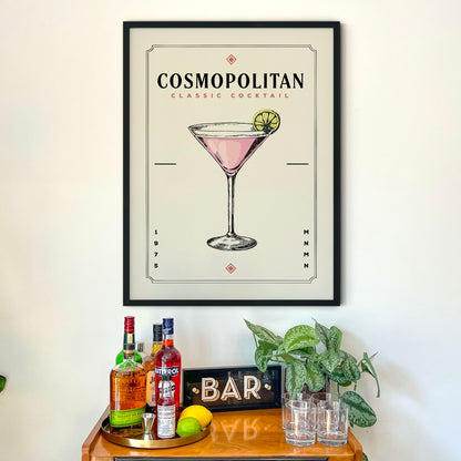 a framed poster of a cocktail glass with a pink drink and a lemon slice, hanging on a wall above a bar counter with various liquor bottles and glasses.