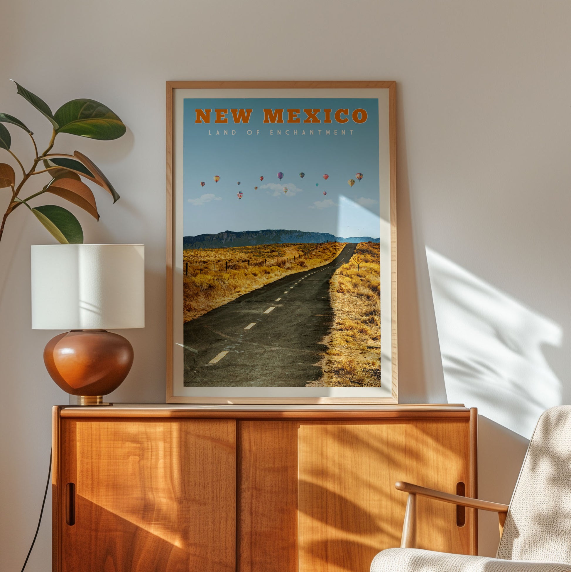 a framed poster of a road leading to a desert landscape with balloons in the sky, hanging on a wall next to a wooden cabinet and a lamp.