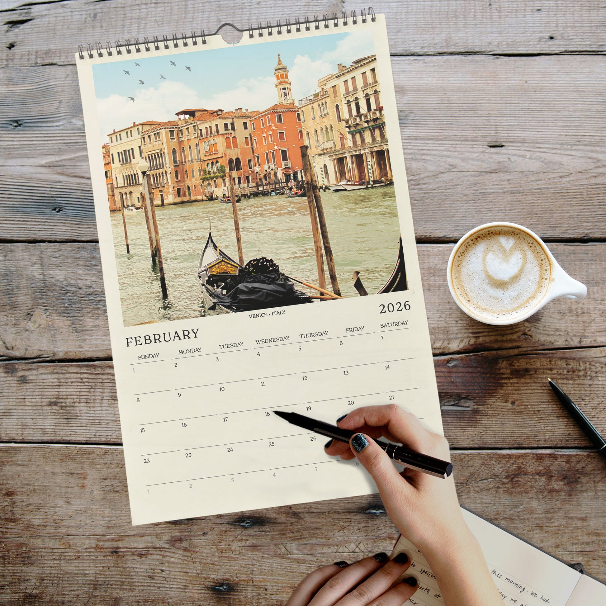 A person's hand is writing in a notebook while holding a pen, with a calendar for February 2026 visible on a wooden surface.
