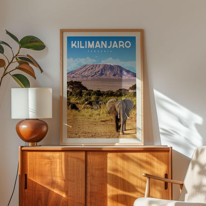 a framed poster of an elephant in a natural setting, with a mountain in the background. The poster is hanging on a wall, and there is a lamp and a plant nearby.