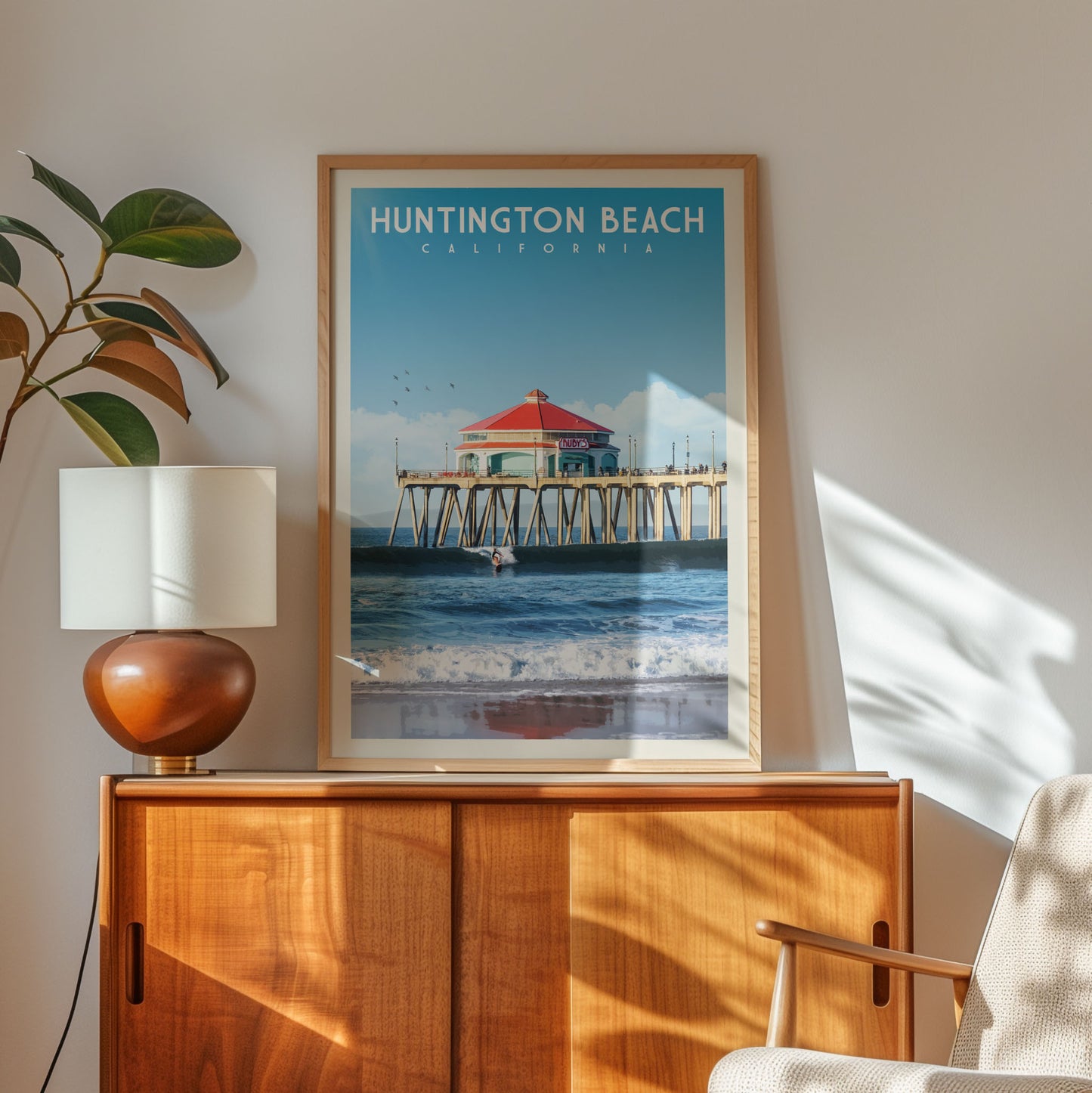 a framed poster of Huntington Beach, California, with a wooden cabinet below it and a plant to the left.