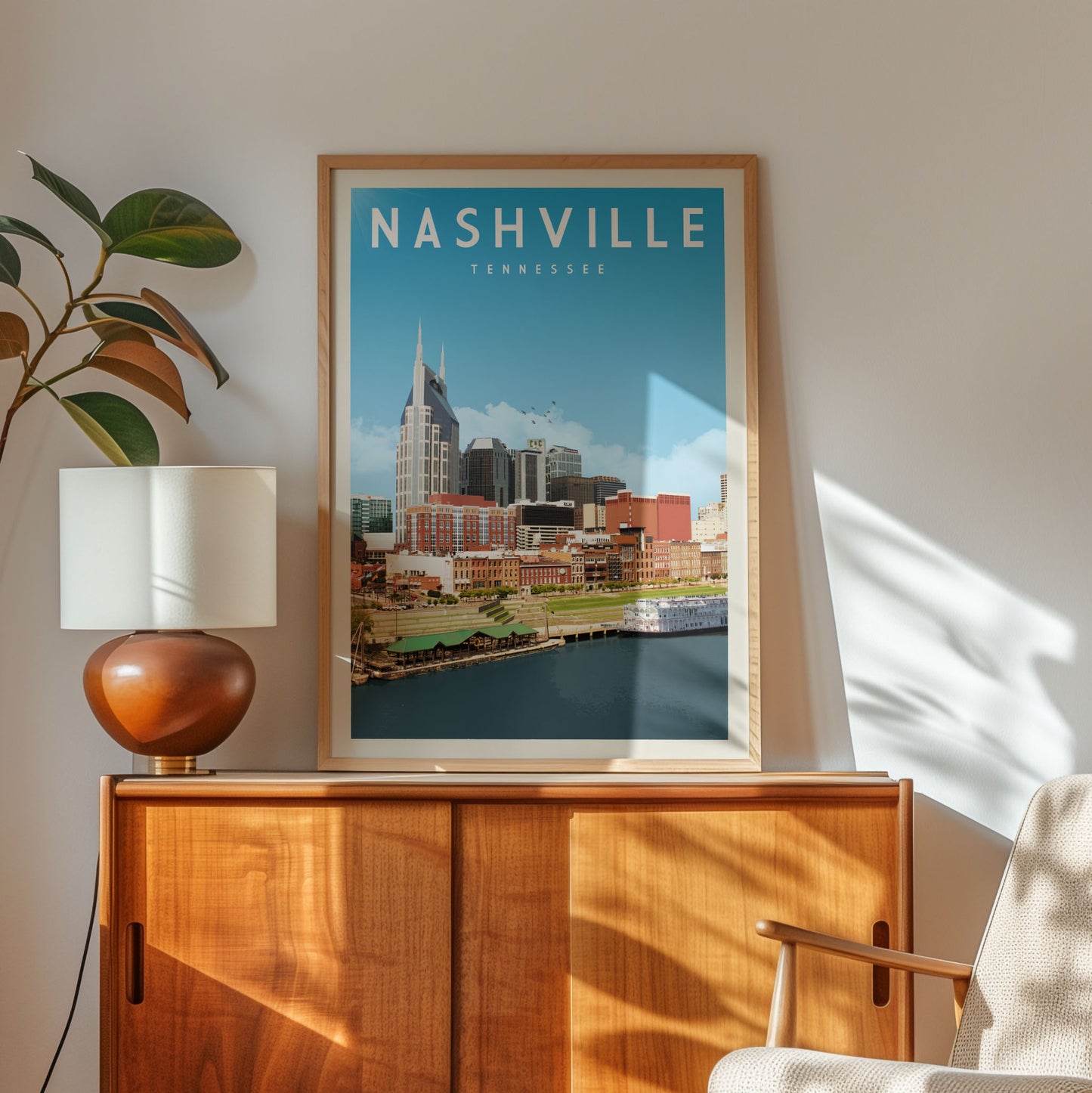 A framed poster of Nashville, Tennessee, featuring a cityscape with a river and buildings, is displayed on a wooden cabinet in a room with a lamp and a plant.